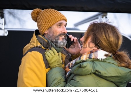 Middle aged couple talking on the phone traveling with camper at winter vacation away in wild. Smiling middle aged man and woman in snowy nature or park together. Family seasonal vacation concept. 