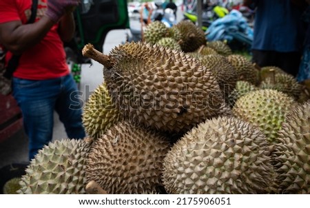 A pile of durians at a tropical fruit market, with blurry background of a man standing nearby the durians. A close-up and selective focus photo of the durian fruit.