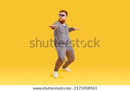 Plus size male model in funny PJs having fun in modern fashion studio. Happy carefree confident fat bearded man in comfortable leopard pajamas and cool glasses dancing isolated on yellow background