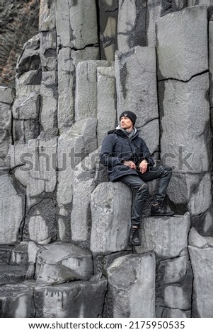 Tourist sitting on beautiful basalt columns. Thoughtful man exploring rocky formation on seashore. Scenic view of gray patterned stones at famous Reynisfjara Beach. Royalty-Free Stock Photo #2175950535