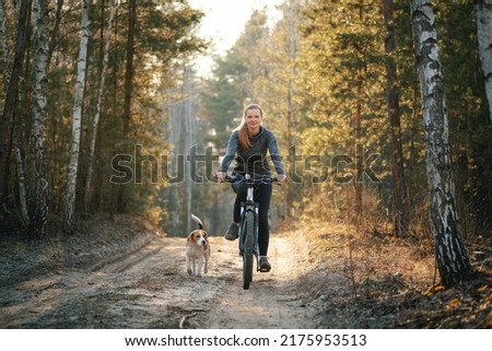 Woman cycling with a dog in the forest. Young woman riding bicycle together with her beagle dog pet running nearby. Traveling with a dog