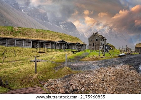 Traditional houses amidst grassy field under majestic Vestrahorn mountain. View of Viking village near Stokksnes against cloudy sky. Historic tourist attraction in volcanic valley during sunset.