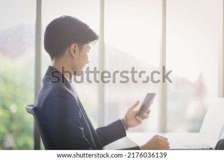 Business Asian man in formal manager looking suit working using smart phone with smiling and confident emotion in modern interior office workplace.