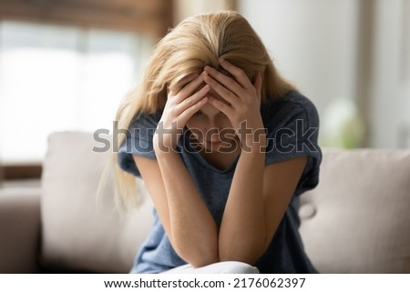 Crazy day. Stressed depressed young adult lady sitting on couch at home with closed eyes hugging head with hands enduring headache, panic attack, feeling bad, tired, shame, sorrow, hopeless, desperate Royalty-Free Stock Photo #2176062397