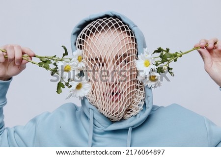 a conceptual studio photograph of a man in a light blue hoodie, a mesh stretched over his face, standing on a light background, holding daisies near his face, looking into the camera