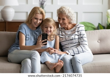 How funny. Happy multi generation family, mature elderly grandma, young millennial mother and cute little daughter grandkid sitting on sofa making call, posing for selfie, watching video on cellphone