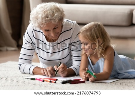 That is how to paint it. Caring mature granny teaching interested granddaughter preschooler to draw sketches on paper sheets, elderly nanny and little girl lying on warm floor indoor coloring pictures