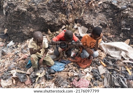 Three little African boys sitting on a landfill suffering from breathing problems, burning eyes, headaches, sore throats and the unbearable smell; Concept of child exploitation and health risks Royalty-Free Stock Photo #2176097799