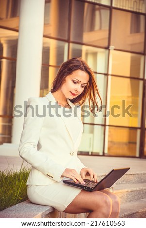 Beautiful young brunette  business woman in white dress working on her laptop outdoors. Heaven sunset glass background behind her. Girl sitting on the grass near modern building
