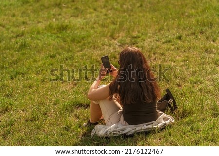 A young woman in light summer clothes chats on a smartphone while sitting on green grass lit by sunlight, a mobile phone for communication with loved ones and work. Back view without face, copy space