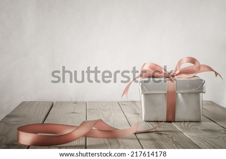 A gift box with closed lid, wrapped in silver paper and tied to a bow with a satin ribbon.  Placed on a weathered old wooden table with copy space behind and above. Cut ribbon remnant to the side.  Royalty-Free Stock Photo #217614178