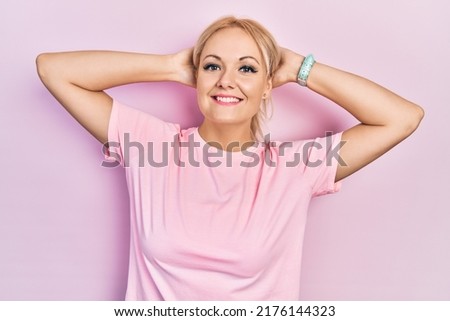 Young blonde woman wearing casual pink t shirt relaxing and stretching, arms and hands behind head and neck smiling happy 