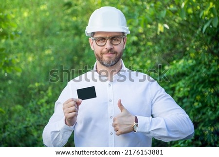 Man foreman in hardhat giving thumbs up showing blank contact card outdoors, copy space