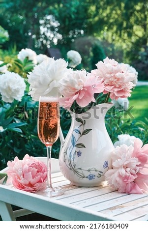Still life with wine glass and rose wine on a wooden tray with a bouquet of peonies and jug on a blurred background in the garden for prepare celebration party in a sunny day. Shallow depth of field