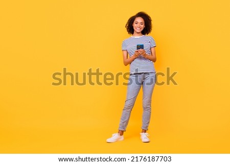 Full length photo of pretty shiny girl dressed striped t-shirt communicating modern gadget empty space isolated yellow color background