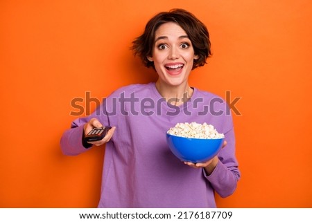 Photo of astonished positive lady hold popcorn plate remote control switch channel isolated on orange color background