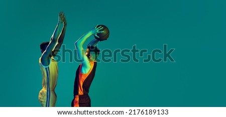 Portrait of two young men, professional basketball players in a jump, throwing ball into basket isolated over blue background in neon light. Concept of sport, team game, action, active lifestyle, ad