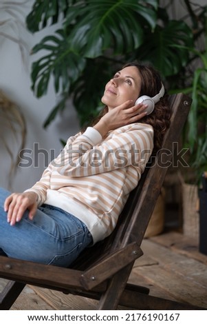 Beautiful woman wearing wireless headphones sits on wooden chair in cozy urban jungle garden, dreaming looks at ceiling, enjoy good meditative music or various podcasts online. Stress free concept. 