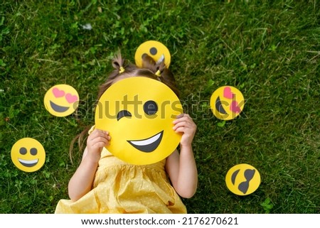 A child hides his face behind a yellow winking emoticon lying on the grass in the park
