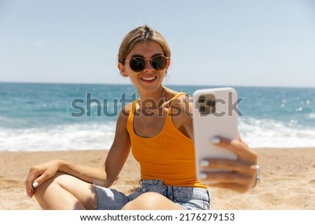 Close up portrait of smiling happy girl in round sunglasses sitting on the beach and making selfie in sunny hot day on sea background 