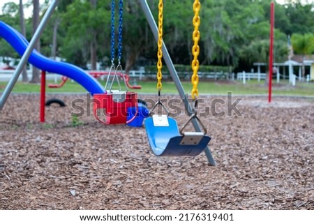 Multiple plastic and rubber swings hanging from chains in a children's park. There's a blue plastic slide in the background with large lush trees and benches. The ground is covered in tree mulch.  Royalty-Free Stock Photo #2176319401