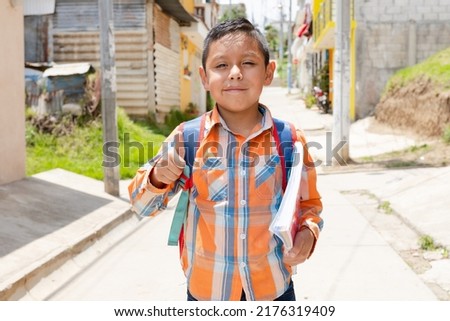 Happy Hispanic boy with thumb up ready to go to school with his backpack and notebooks - Latin boy on his way to school - Happy boy in the village Royalty-Free Stock Photo #2176319409