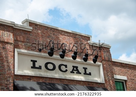 A cream colored informational sign with the word local in black lettering is affixed to a vintage red and brown colored brick building. There are multiple black barn lights hanging over the signage. 
