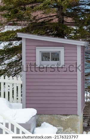 A small purple and grey wooden building with a small window. The outdoor toilet, outhouse, has a black roof.The building has trees in the background with a white picket fence in the foreground.