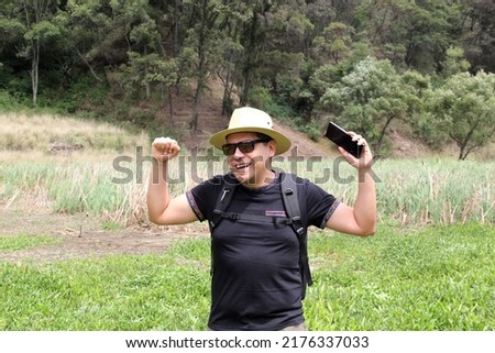 Latin adult man with hat, shorts and sunglasses uses his cell phone in the middle of the forest outdoors to make calls, receive messages, locate on the map because he is lost