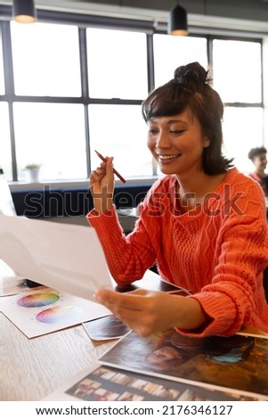 Smiling young creative asian businesswoman with photograph at desk in office. Unaltered, creative business, workplace, photography themes.