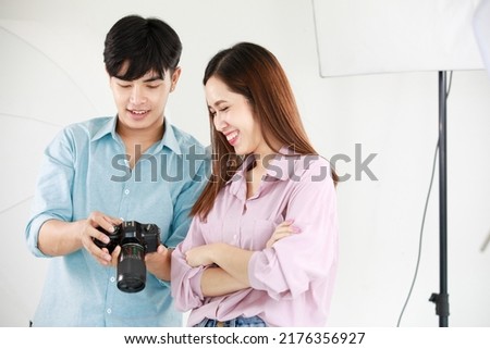 Asian young beautiful long brown hair female model and handsome male photographer holding full frame dslr camera in hands stand smiling talking together checking shooting image from screen in studio.