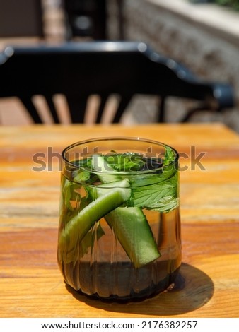 Cucumber and mint leaves infused water, cocktail, lemonade in glass in cafe. Summer iced cold drink with cucumber, ice and mint on orange wooden table outside background, copy space, vertical, sunny