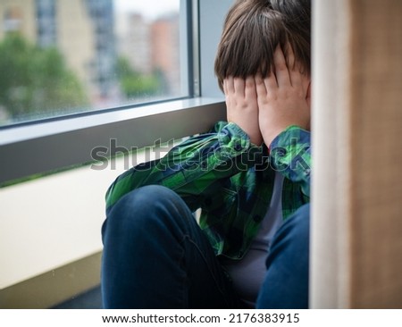 A frustrated, sad and lonely teenager of 12 years old is sitting on the windowsill of the house behind a curtain, covering his face with his hands. Children's social psychology and emotions.