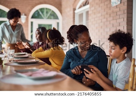 Happy black woman and her grandson having fun while using cell phone at dining table during family lunch on patio.