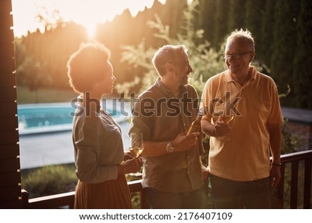 Happy senior man enjoying in conversation with his mature son and African American daughter in law while having a drink on patio at sunset.