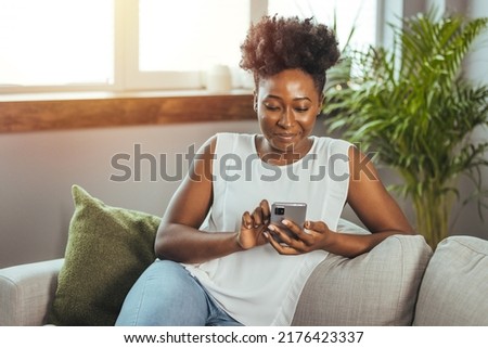 Happy woman text messaging on smart phone at home. Smiling african woman daydreaming and holding mobile phone with copy space. Happy beautiful black girl thinking while using smart phone.