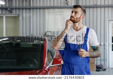 Minded young professional technician car mechanic repairman man wear denim blue overalls white t-shirt hold wrench key work in light modern vehicle repair shop workshop indoors. Tattoo translate fun Royalty-Free Stock Photo #2176430803