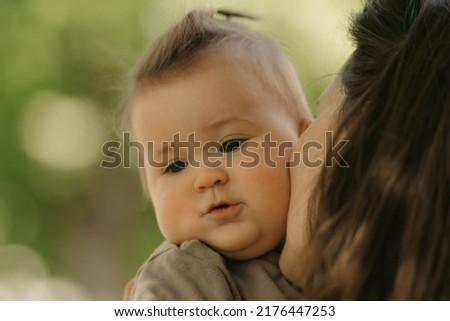A close portrait of a 7-month girl who is been kissing by her mother in the park. A mom is holding in her arms her infant daughter in the woodland. 