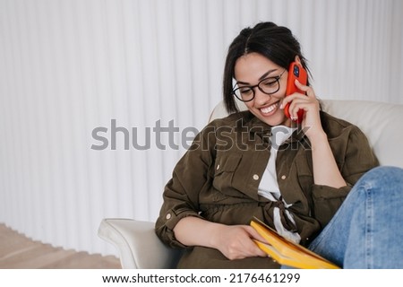 Adorable African american woman in glasses holding diary, speaking by phone, looking shy down, toothy smiling, good news received. Great deal. Entrepreneur satisfied by month profit. Success concept.