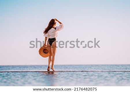 A young attractive woman dressed in a white shirt and bikini, holding a hat in her hand, looks towards the sunset and runs through the water with her hair flying. Vacation at the sea in the summer.