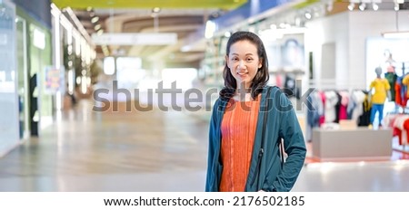 Woman wearing orange shirt and blue jacket put her hand in the pocket and smile with camera in shopping mall. 