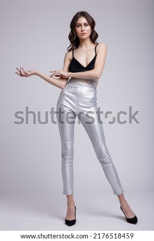 High fashion photo of a beautiful elegant young  woman in a pretty silver trousers, pants, black top posing on white background. Slim figure, studio shot. Brunette, long hair, curls Royalty-Free Stock Photo #2176518459