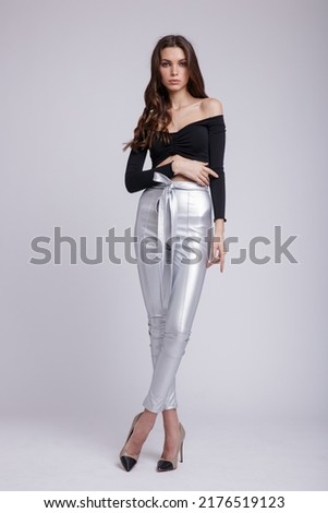 High fashion photo of a beautiful elegant young  woman in a pretty silver trousers, pants, black top with sleeves posing on white background. Slim figure, studio shot. Brunette, long hair Royalty-Free Stock Photo #2176519123