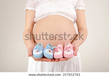 Close up on pregnant belly with pink and blue new born shoes.. Woman expecting a baby dressed in white holding girl and boy baby boots.