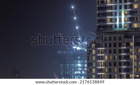 Tall buildings under construction and cranes in downtown night timelapse. Work progress at construction site of new towers behing skyscraper with glowing windows