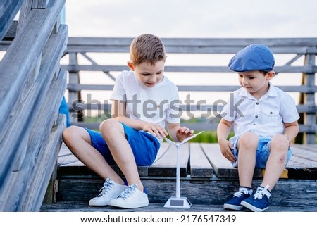 Two happy brothers children learning how working model of a wind turbine while sitting at wooden pier in lake summer vacation nature. Renewable energy, kids, technology, science and people concept.