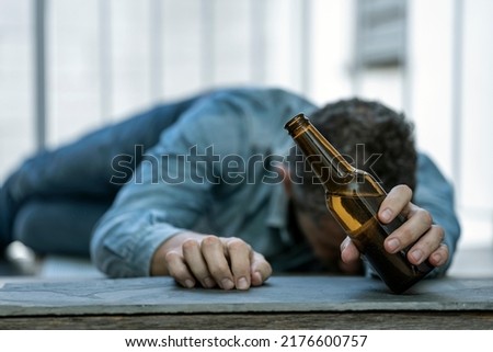 DRUNK MAN LYING ON THE FLOOR ASLEEP WITH A BOTTLE OF BEER IN HIS HAND. ALCOHOL CONSUMPTION ADDICTION. ALCOHOLISM CONCEPT. FOCUS SELECTED. Royalty-Free Stock Photo #2176600757