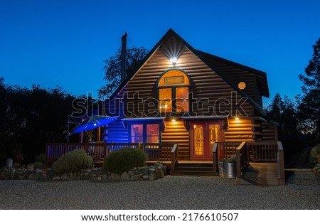 Exterior of rustic cabin home at dusk with lights turned on.  A patio umbrella is open with lights on and a blue accent light below. The inside of the home can be clearly seen. Taken March 2021.