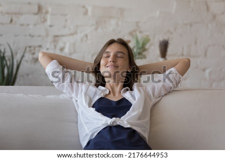 Calm young 20s freckled woman relaxing on comfortable couch at home, breath fresh ventilated conditioned air inside modern living room. Enjoy carefree leisure, day off, climate control, rest concept Royalty-Free Stock Photo #2176644953