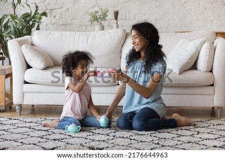 African mother and little daughter holding plastic toy teacups enjoy tea ceremony play together sit on carpet in warm living room. Dishware shop advertisement, weekend playtime with children concept Royalty-Free Stock Photo #2176644963